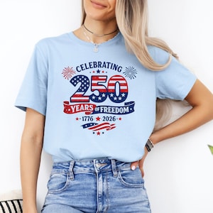 May include: A light blue t-shirt featuring a graphic design with the text CELEBRATING 250 YEARS OF FREEDOM 1776-2026. The design includes red, white, and blue colors, star patterns, firework illustrations, and a waving flag motif across the chest.