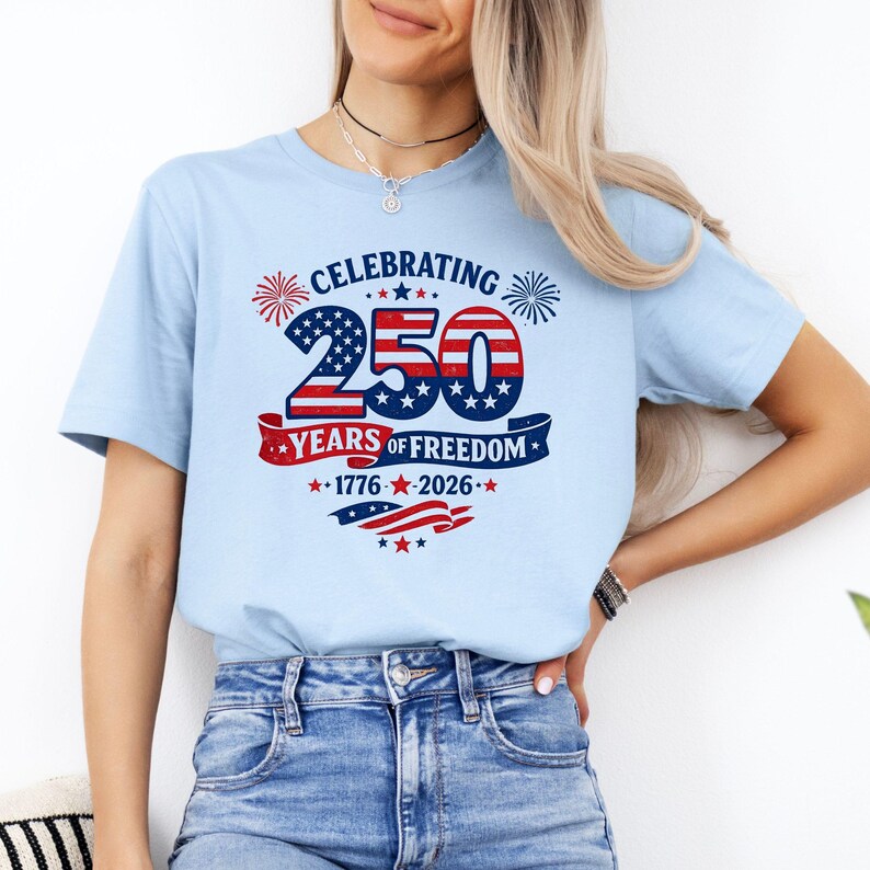 May include: A light blue t-shirt featuring a graphic design with the text CELEBRATING 250 YEARS OF FREEDOM 1776-2026. The design includes red, white, and blue colors, star patterns, firework illustrations, and a waving flag motif across the chest.