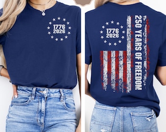 May include: Navy blue t-shirt with a distressed American flag design on the back, featuring red and white stripes and the text "250 YEARS OF FREEDOM." The front displays a circle of stars with the dates "1776 2026."