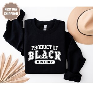 Op de afbeelding: Zwarte sweatshirt met de witte tekst "PRODUCT OF BLACK HISTORY". De sweatshirt wordt gepresenteerd op een witte achtergrond, met een beige hoed en decoratieve bladeren.