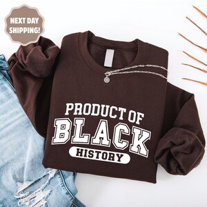 Op de afbeelding: Een donkerbruine sweatshirt met de witte tekst "PRODUCT OF BLACK HISTORY". Een zilveren ketting met een kleine hanger ligt op de sweatshirt. De sweatshirt is op een blauwe spijkerbroek gevouwen.