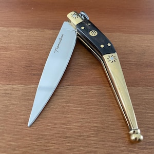 May include: A partially open folding knife with a silver blade and a dark wood handle accented with gold-colored metal. The blade is engraved with the word "Tramontina". The knife is resting on a wooden surface.