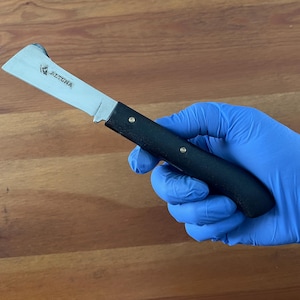 May include: A silver and black knife with the word "ALTUNA" printed on the blade. The knife has a black handle and is held by a person wearing a blue glove. The background is a wooden surface.
