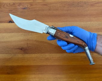 May include: A stainless steel knife with a curved blade and a wooden handle. The handle is a dark brown color with silver accents. The knife is held by a person wearing a blue glove.