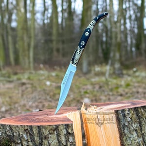May include: A decorative knife with a curved handle and a silver blade is embedded in a tree stump. The handle is black with silver floral accents. The knife is positioned in the center of the image, with a blurred forest background.