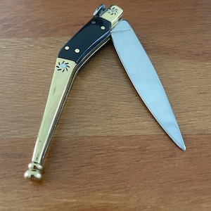 May include: A small, black-handled pocket knife with a brass handle and a silver blade. The blade is open and the knife is lying on a wooden surface.