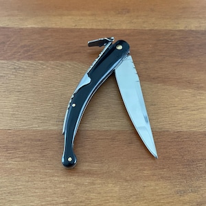 May include: A black handled pocket knife with a curved blade and a gold accent. The knife is open and the blade is visible.