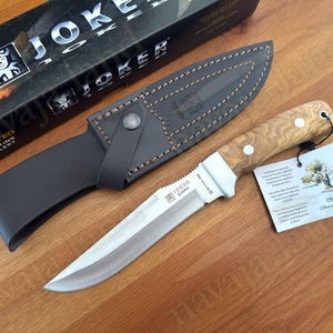 May include: A hunting knife with a wooden handle and a leather sheath. The knife blade is silver, and the handle has a light brown wood grain pattern. The sheath is black with a button closure. The box says "Joker".