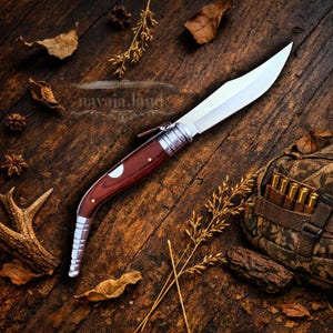 May include: A handcrafted folding knife with a polished steel blade and a dark wood handle. The handle is accented with brass rivets and a silver bolster. The knife is displayed on a rustic wooden surface with natural elements.