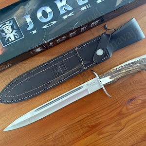 May include: A hunting knife with a polished steel blade and a textured antler handle, resting on a wooden surface. The knife is accompanied by a dark brown leather sheath. The box in the background has the word "JOKER" on it.