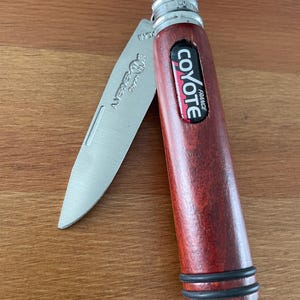 May include: A partially open pocket knife with a reddish-brown wooden handle. The handle features the words "Coyote France". The silver blade is partially extended and has the word "INOX" printed on it.