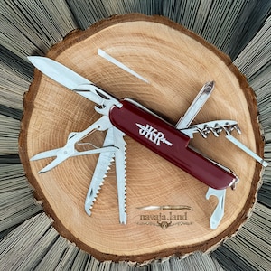 May include: A multi-tool pocket knife with a deep red handle featuring the silver text JKR and a small knife icon. The tool includes a main blade, scissors, a saw, a corkscrew, a bottle opener, and other metal implements extended outward.
