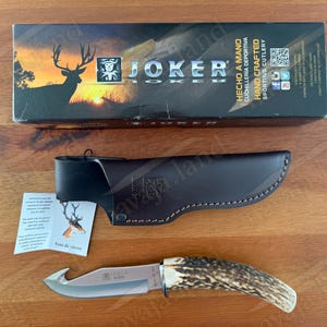 May include: A hunting knife with a brown antler handle and a stainless steel blade, accompanied by a leather sheath and a box. The box features a deer silhouette and the text "JOKER". The knife is designed for sporting use.