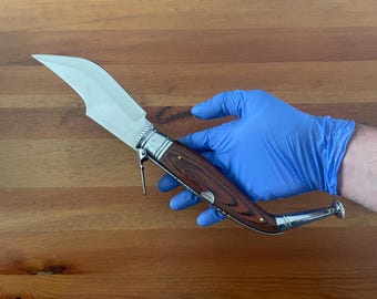 May include: A stainless steel and wood-handled knife with a curved blade. The handle is a dark wood with brass rivets and a silver bolster. The knife is held by a person wearing a blue glove. The knife is on a wooden surface.