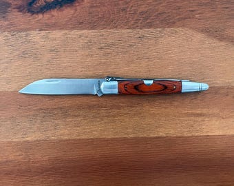 May include: A silver folding pocket knife with a wooden handle. The handle is a dark brown wood with a reddish hue. The blade is closed and the knife is lying on a wooden surface.