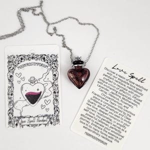 May include: A silver chain necklace with a glass vial pendant shaped like a heart. The vial is filled with a dark purple liquid and has a silver cap. The necklace is accompanied by a card with the text "Love Spell" and a description of the spell's ingredients and purpose.