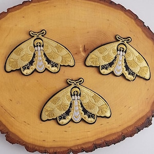 Puede incluir: Tres parches de polilla bordados en dorado con detalles en negro y blanco. Las polillas tienen un diseño de luna creciente y estrella en sus alas.