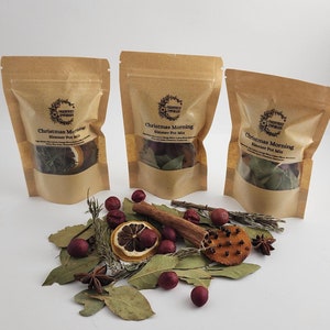 Könnte beinhalten: Drei braune Papiertüten mit der Aufschrift "Christmas Morning Simmer Pot Mix", gefüllt mit getrockneten Kräutern und Gewürzen. Vorne sind Lorbeerblätter, Zimtstangen, getrocknete Orangenscheiben und Sternanis angeordnet.