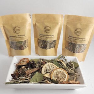 以下が含まれることがあります： 3つの茶色の紙袋に「Immunity Strength」と「Smudge Pot Manifestation Kit」の文字が印刷されています。袋には乾燥ハーブとスパイスが入っています。白いボウルには、乾燥ハーブ、スパイス、柑橘類の皮が入っています。