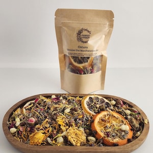 May include: A brown paper bag with a white label that says "Ostara Simmer Pot Manifestation Kit" and a list of ingredients. The bag is filled with dried herbs, flowers, and citrus peels. The bag is sitting on a wooden tray with a similar mix of dried herbs, flowers, and citrus peels.