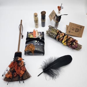 May include: A ritual kit featuring a small broom decorated with orange dried flowers and mini pumpkins, a black feather, a bundle of dried herbs wrapped in black string, two glass vials, and a rectangular box wrapped in black paper with moon phases.