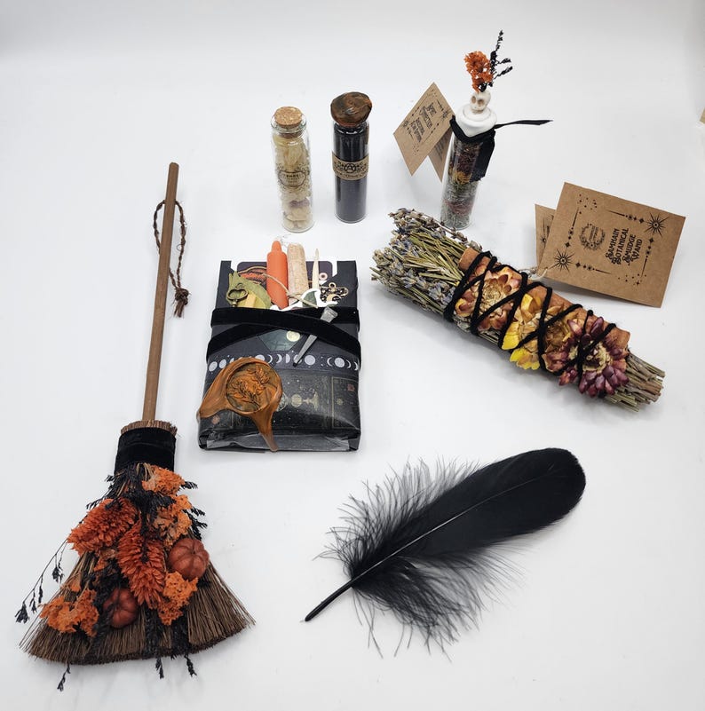 May include: A ritual kit featuring a small broom decorated with orange dried flowers and mini pumpkins, a black feather, a bundle of dried herbs wrapped in black string, two glass vials, and a rectangular box wrapped in black paper with moon phases.