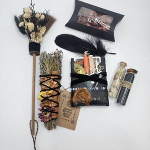 May include: A collection of ritual items including a small broom with dried flowers, a black box labeled MINI PUMPKINS, a black feather, a botanical smudge wand with a tag reading SAMHAIN BOTANICAL SMUDGE WAND, a wrapped package, and two glass vials.
