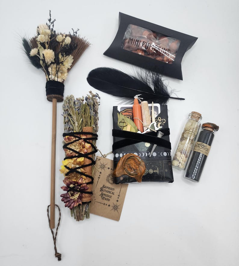 May include: A collection of ritual items including a small broom with dried flowers, a black box labeled MINI PUMPKINS, a black feather, a botanical smudge wand with a tag reading SAMHAIN BOTANICAL SMUDGE WAND, a wrapped package, and two glass vials.