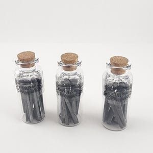 May include: Three clear glass bottles with cork stoppers, each containing gray coffin nails. The bottles have labels with the text "Coffin Nails" in a decorative font. The nails are arranged vertically inside the bottles.