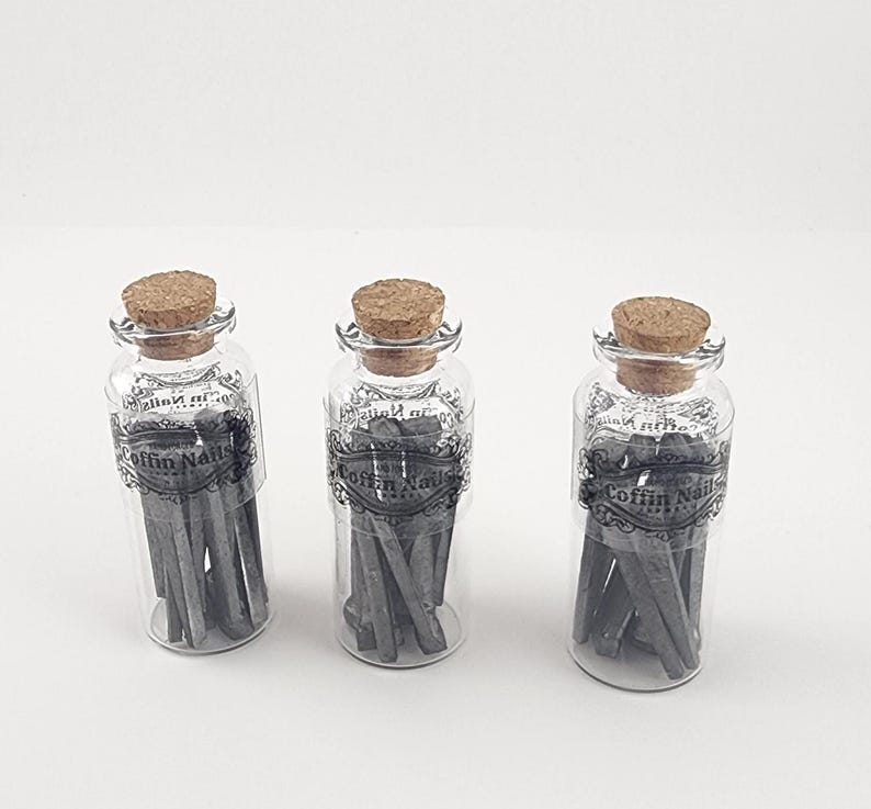 May include: Three clear glass bottles with cork stoppers, each containing gray coffin nails. The bottles have labels with the text "Coffin Nails" in a decorative font. The nails are arranged vertically inside the bottles.