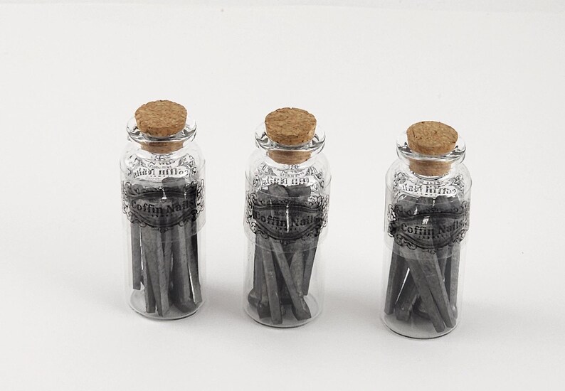 May include: Three clear glass bottles with cork stoppers, each filled with gray coffin nails. The bottles have decorative labels with the text "Coffin Nails". The background is plain white.