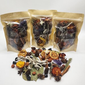 May include: Three clear stand-up pouches filled with dried botanicals, pinecones, and crystals, labeled with text. In front, a loose collection of dried orange slices, white sage bundles, cinnamon sticks, star anise, and small pinecones.