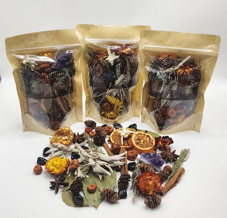 May include: Three clear stand-up pouches filled with dried botanicals, pinecones, and crystals, labeled with text. In front, a loose collection of dried orange slices, white sage bundles, cinnamon sticks, star anise, and small pinecones.
