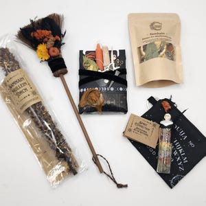 May include: A Samhain ritual kit featuring a mullein torch in plastic packaging, a small broom with dried flowers, a black box with moon phases containing small items, a pouch of dried herbs, a small card, and a glass vial on a ouija board pouch.