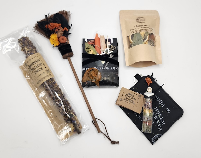 May include: A Samhain ritual kit featuring a mullein torch in plastic packaging, a small broom with dried flowers, a black box with moon phases containing small items, a pouch of dried herbs, a small card, and a glass vial on a ouija board pouch.