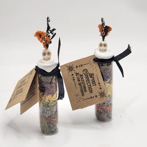 May include: Two clear glass vials filled with layers of dried herbs and flowers, topped with a white skull-shaped stopper and orange dried sprigs. Each vial features a black ribbon and a tan paper tag printed with Spirit Connection Ancestral Altar Offering.