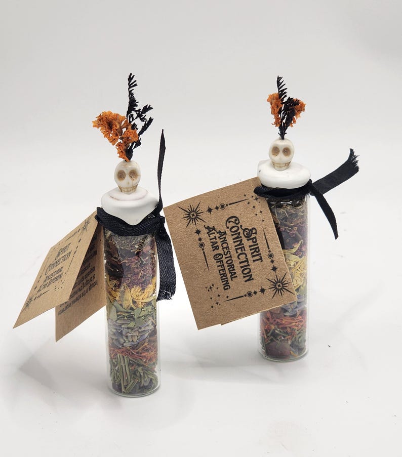 May include: Two clear glass vials filled with layers of dried herbs and flowers, topped with a white skull-shaped stopper and orange dried sprigs. Each vial features a black ribbon and a tan paper tag printed with Spirit Connection Ancestral Altar Offering.