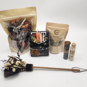 May include: A collection of items including a large pouch of dried botanicals, a black box containing an orange candle and herbs, a smaller pouch labeled Samhain, two glass vials with cork stoppers, and a small broom with dried white flowers.