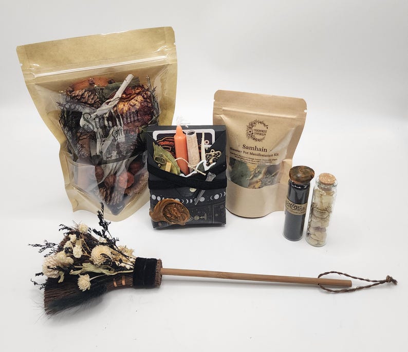 May include: A collection of items including a large pouch of dried botanicals, a black box containing an orange candle and herbs, a smaller pouch labeled Samhain, two glass vials with cork stoppers, and a small broom with dried white flowers.