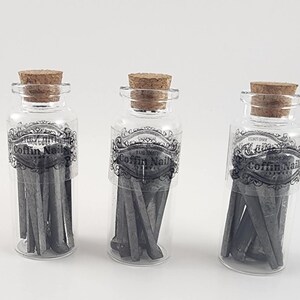 May include: Three clear glass bottles with cork stoppers, each containing dark gray coffin nails. The bottles have a black and white label that reads "COFFIN NAILS". The nails are visible through the transparent glass.