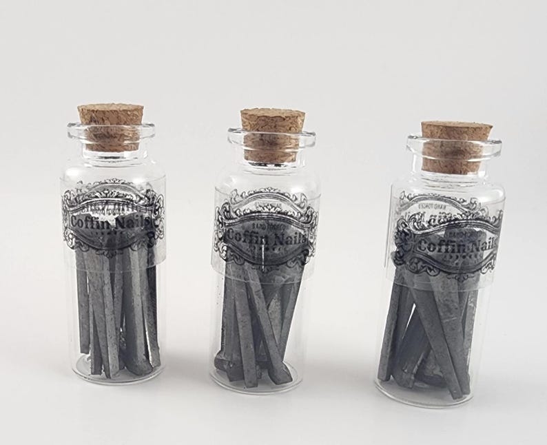 May include: Three clear glass bottles with cork stoppers, each containing dark gray coffin nails. The bottles have a black and white label that reads "COFFIN NAILS". The nails are visible through the transparent glass.