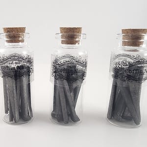 May include: Three clear glass bottles with cork stoppers, each containing gray coffin nails. The bottles have decorative labels with the text "Coffin Nails". The nails are arranged inside the bottles, creating a unique display.