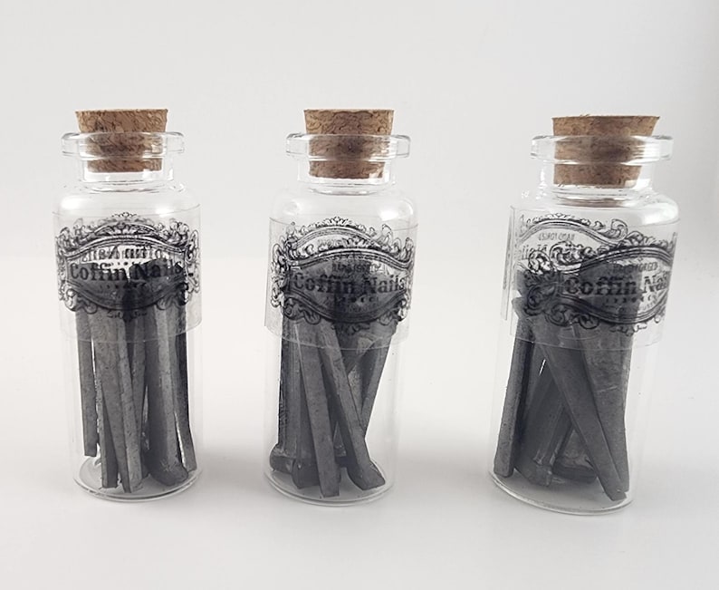 May include: Three clear glass bottles with cork stoppers, each containing gray coffin nails. The bottles have decorative labels with the text "Coffin Nails". The nails are arranged inside the bottles, creating a unique display.