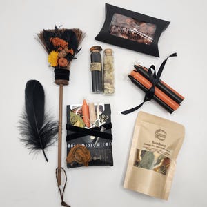 May include: A ritual kit containing a small broom decorated with orange and yellow flowers, two glass vials with cork stoppers, a black box labeled Mini Pumpkins, a bundle of black and orange candles, a black pouch with charms, and a Samhain simmer pot kit.