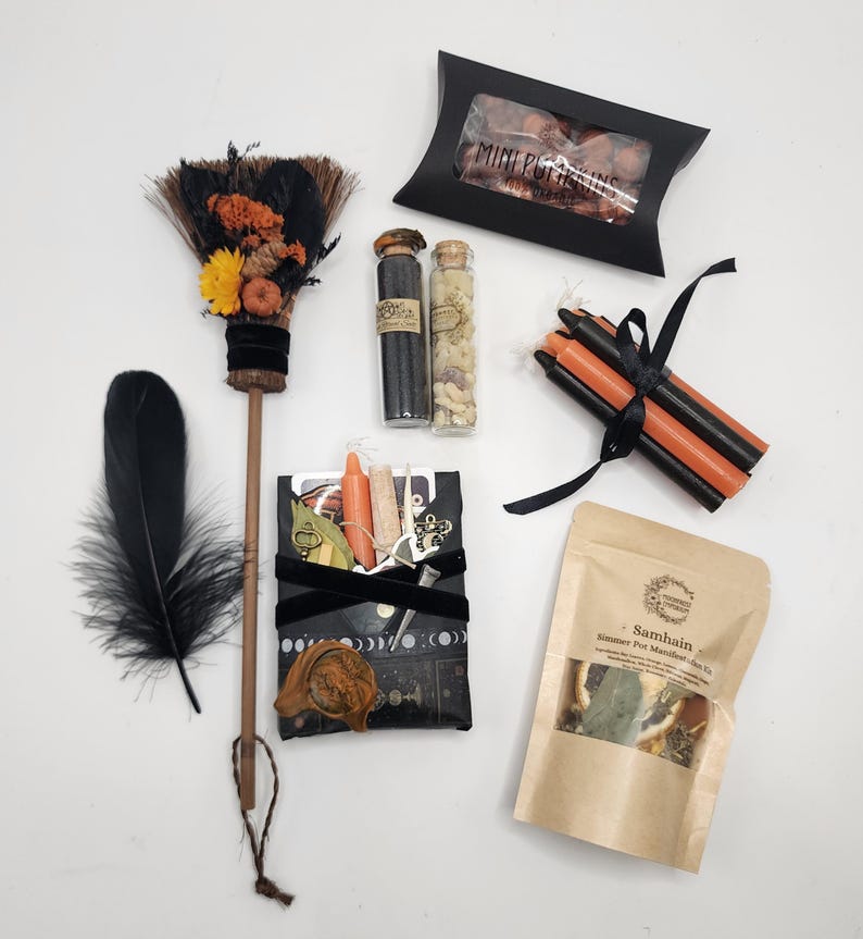 May include: A ritual kit containing a small broom decorated with orange and yellow flowers, two glass vials with cork stoppers, a black box labeled Mini Pumpkins, a bundle of black and orange candles, a black pouch with charms, and a Samhain simmer pot kit.