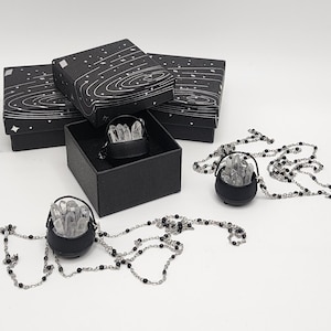Puede incluir: Collares con colgantes de caldero negro con piedras de cuarzo cristalino transparente. Los collares tienen una cadena de cuentas negras. Los colgantes de caldero se exhiben en cajas negras con un diseño de noche estrellada.