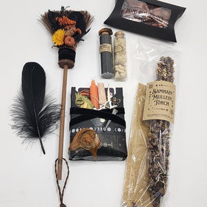 May include: A collection of items including a small broom decorated with dried flowers, two glass vials with contents, a black feather, a black box labeled Mini Pumpkins, and a Samhain Mullein Torch in a clear bag, plus a wrapped package with a wax seal.