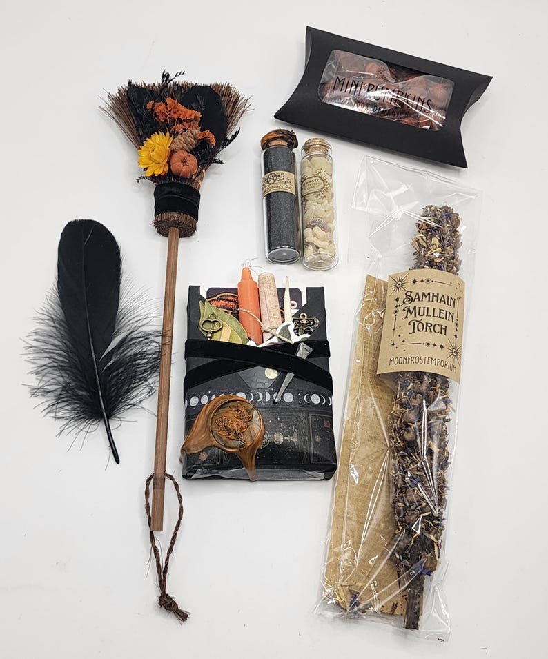 May include: A collection of items including a small broom decorated with dried flowers, two glass vials with contents, a black feather, a black box labeled Mini Pumpkins, and a Samhain Mullein Torch in a clear bag, plus a wrapped package with a wax seal.