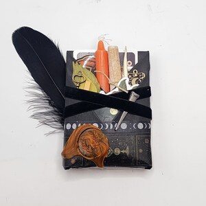 May include: A rectangular black package wrapped with black ribbon, featuring a moon phase pattern and a floral wax seal. It includes a black feather, an orange candle, a cork vial, a small metal key, a metal nail, and a gold-toned charm.