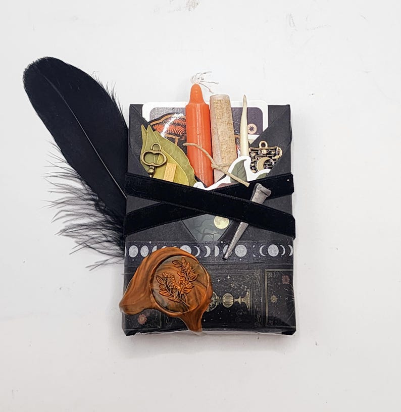 May include: A rectangular black package wrapped with black ribbon, featuring a moon phase pattern and a floral wax seal. It includes a black feather, an orange candle, a cork vial, a small metal key, a metal nail, and a gold-toned charm.